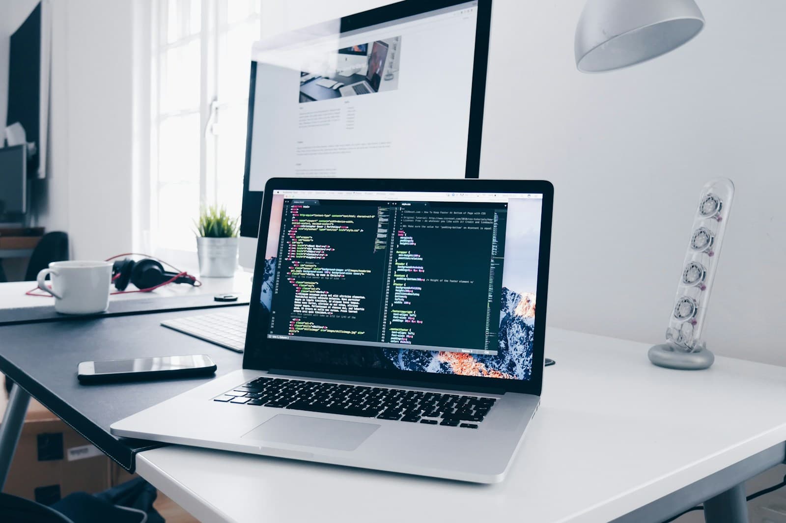 Laptop on a desk showing HTML and CSS source code, with a website design on a monitor behind it