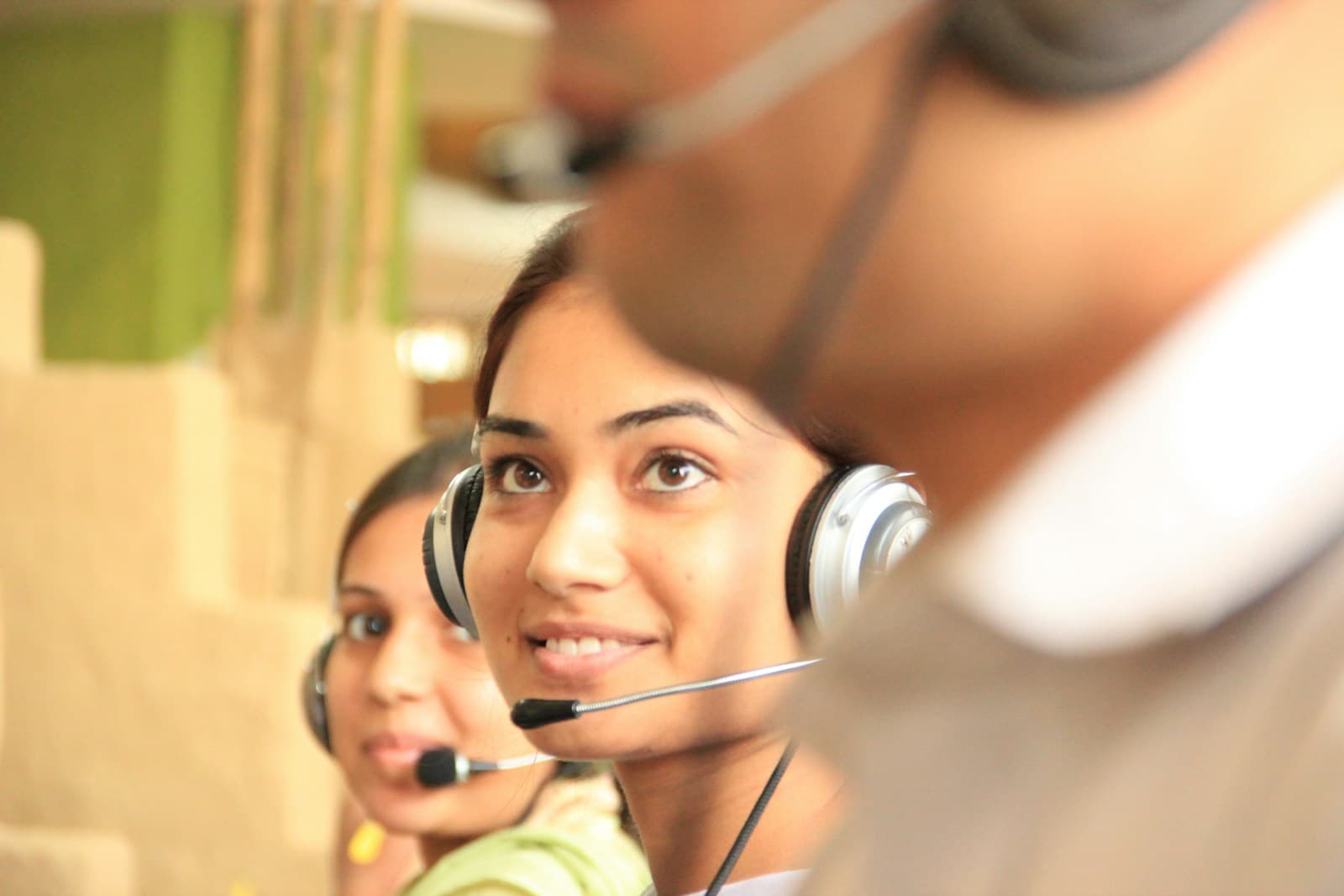 A smiling customer support agent wearing a headset at her desk, with colleagues in headsets behind her