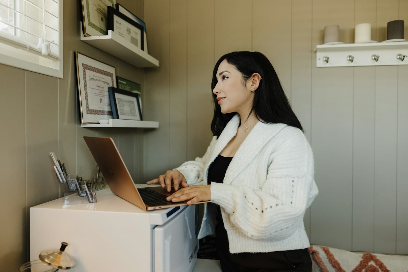 A small business owner in a white cardigan working on a laptop in her studio, framed certificates on the wall behind her