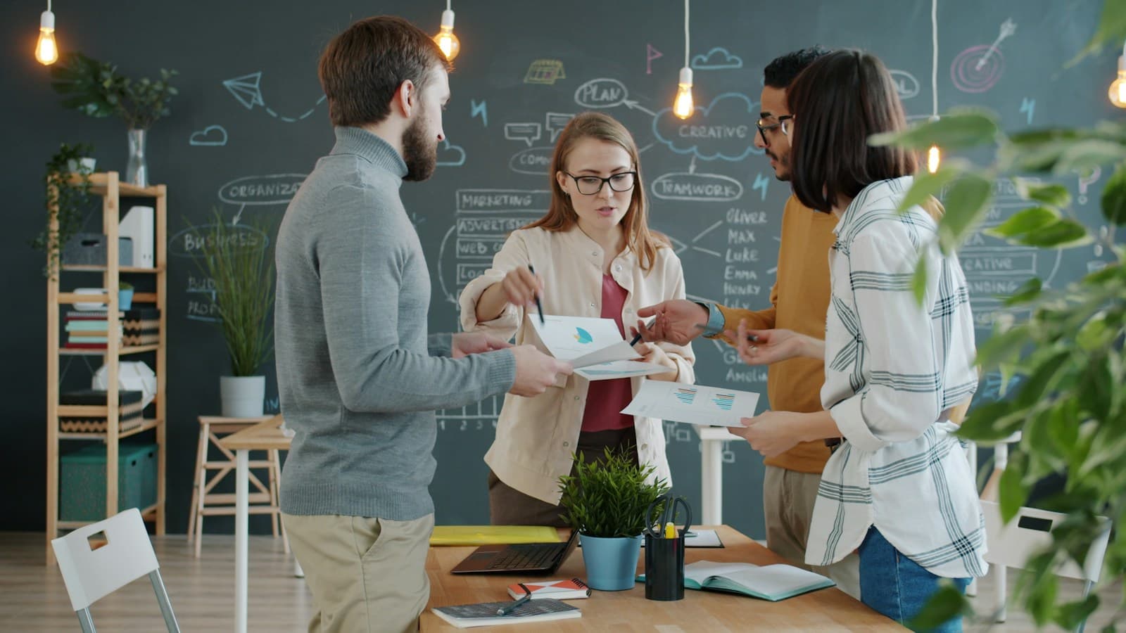 Four colleagues standing around a desk comparing printed charts, with a chalkboard of planning diagrams behind them