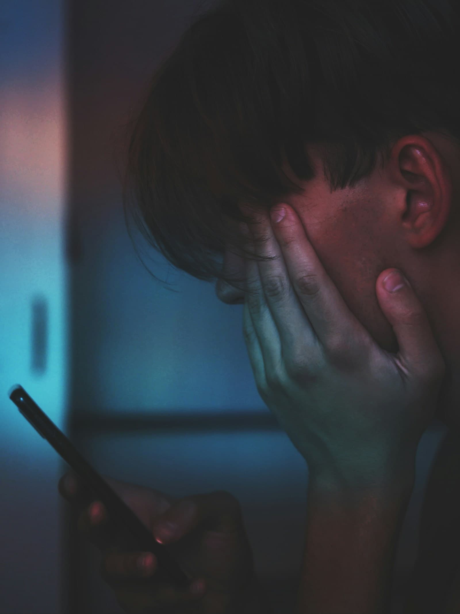 A man in dim light holding his phone with one hand pressed to his face in frustration