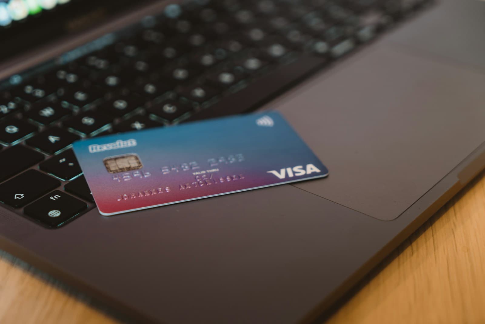 A blue and red Visa debit card resting on the keyboard of a dark laptop on a wooden desk