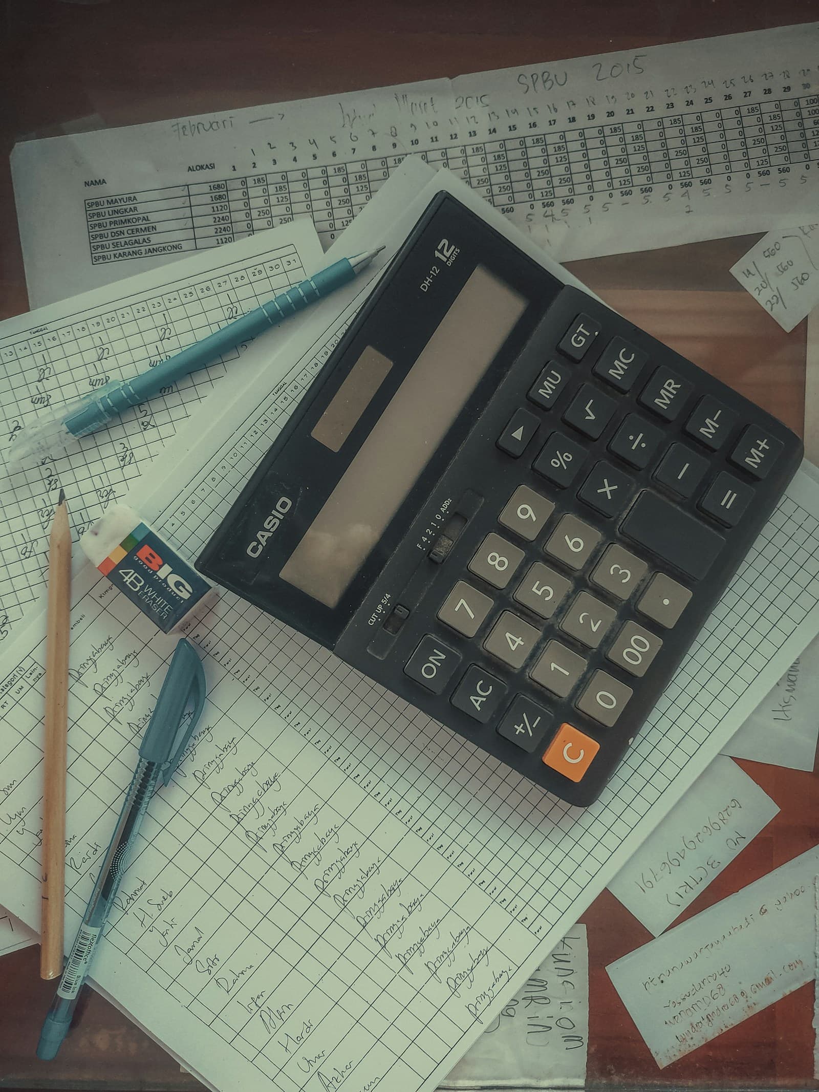 A black desk calculator resting on handwritten ledger sheets and printed spreadsheets, with a pen and pencil beside it