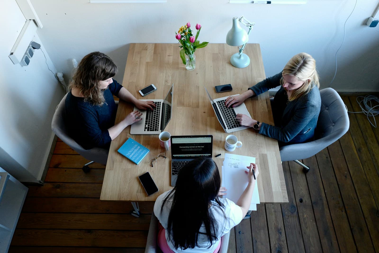 Three women working on laptops around a wooden table in a small office, seen from above