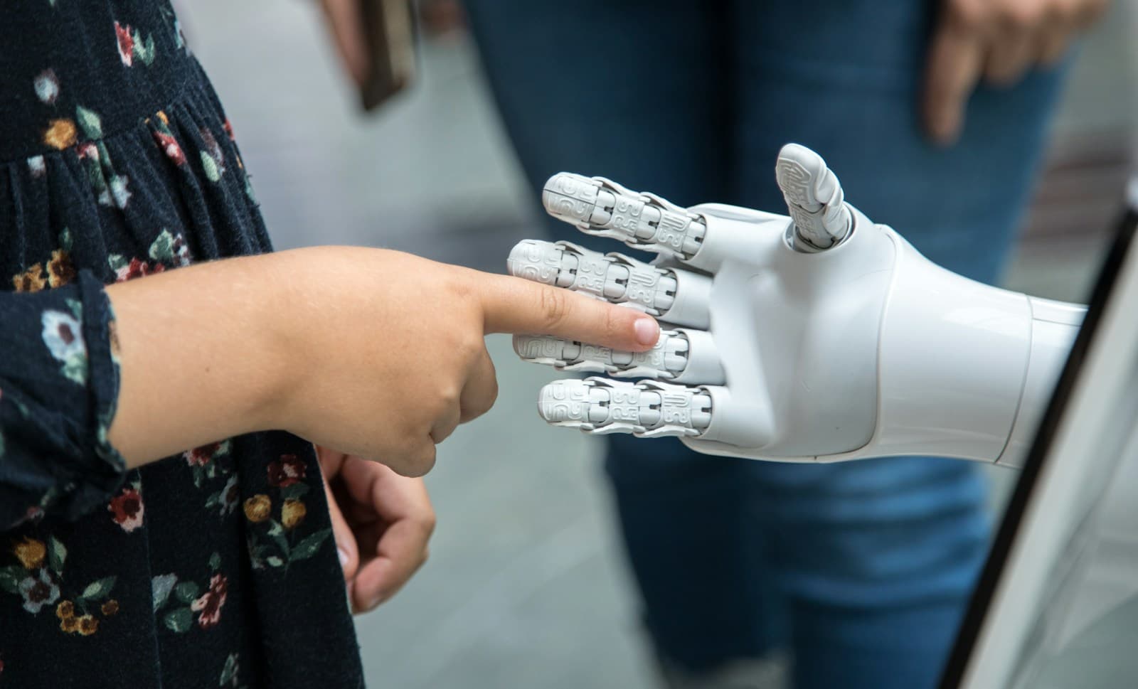 A child's finger touching the fingertip of a white robotic hand, with people standing in the background