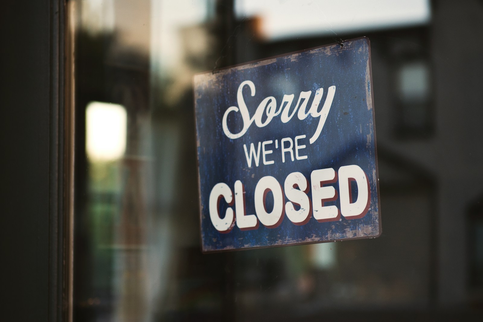 A blue Sorry We're Closed sign hanging inside a glass shop door, with the street blurred behind it