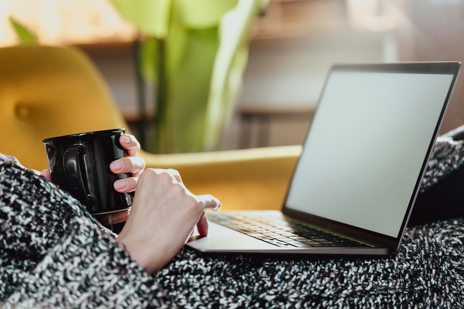 A person wrapped in a knitted blanket holds a black mug in one hand and uses a laptop trackpad with the other