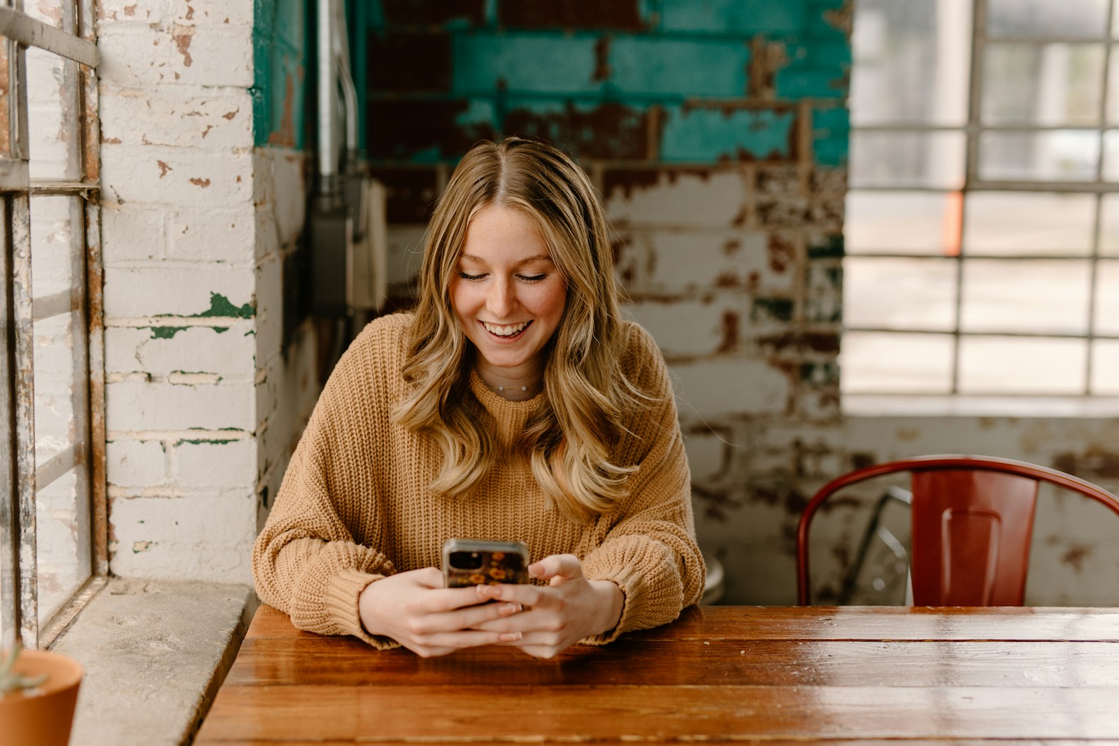 A smiling woman in a tan knit sweater looks at her phone at a wooden table beside an old painted brick wall
