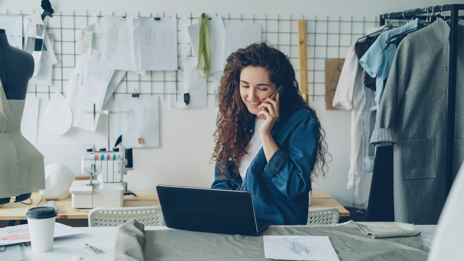 A smiling dressmaker talks on her phone while working on a laptop in a sewing studio with clothing racks