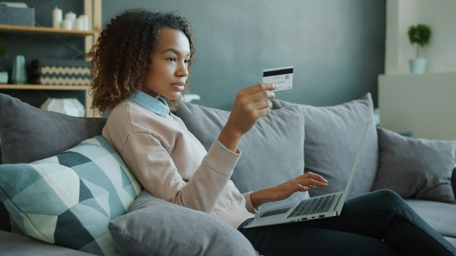 A woman with curly hair sits on a grey sofa holding a bank card while typing on a laptop resting on her lap