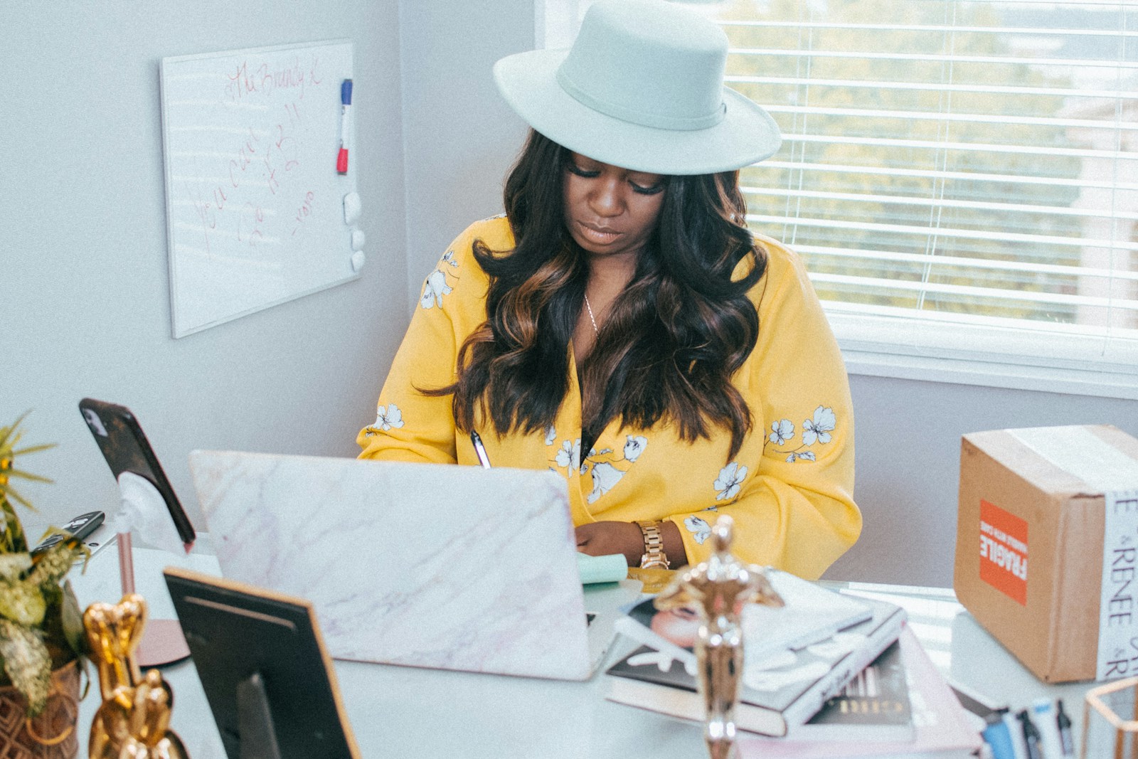 A woman in a yellow floral blouse and pale hat works at a laptop beside a parcel marked fragile