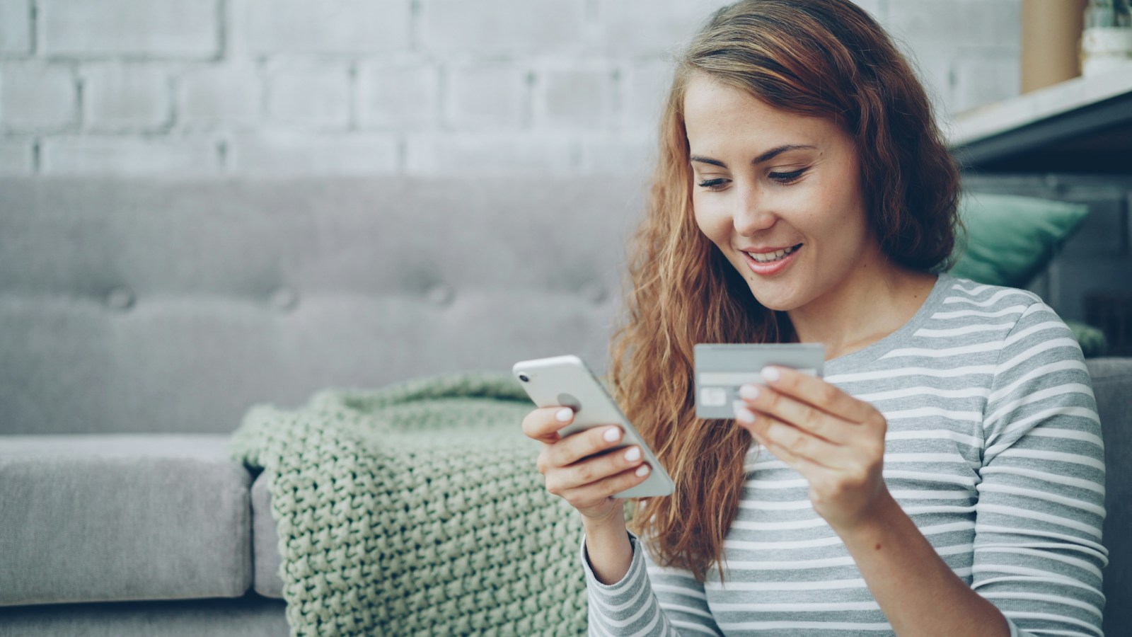 A smiling woman sitting by a grey sofa holds a smartphone in one hand and a bank card in the other while shopping online