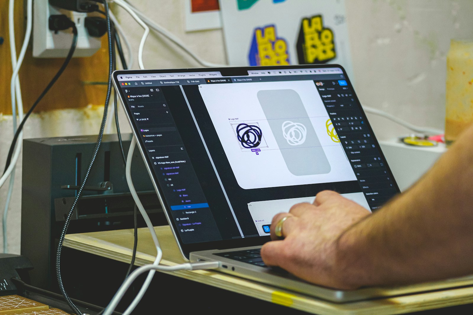A hand on a laptop trackpad while a design app on screen shows a circular logo on a grey artboard