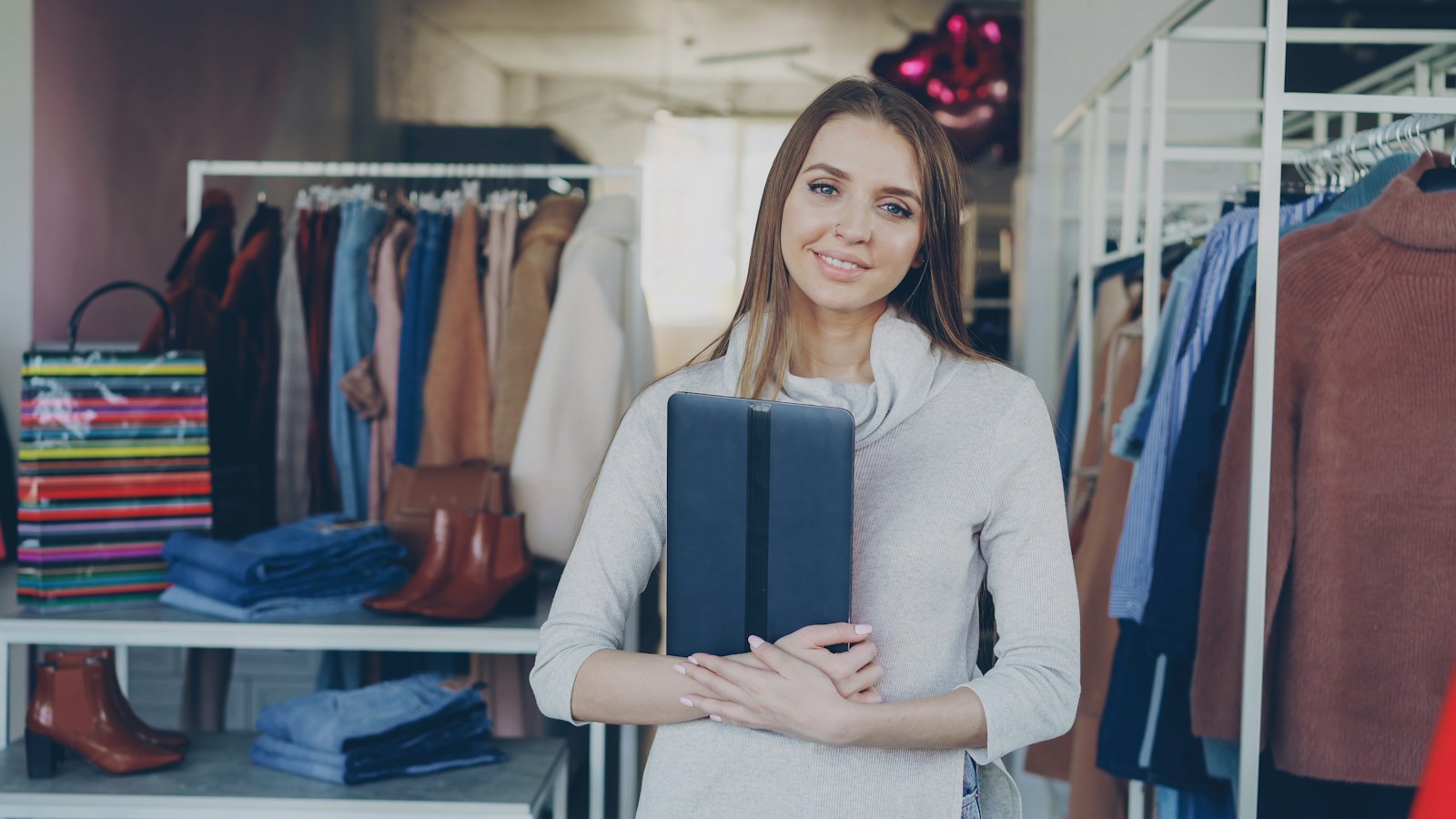 A smiling boutique owner holds a closed tablet among clothing racks, folded jeans and brown boots in her shop