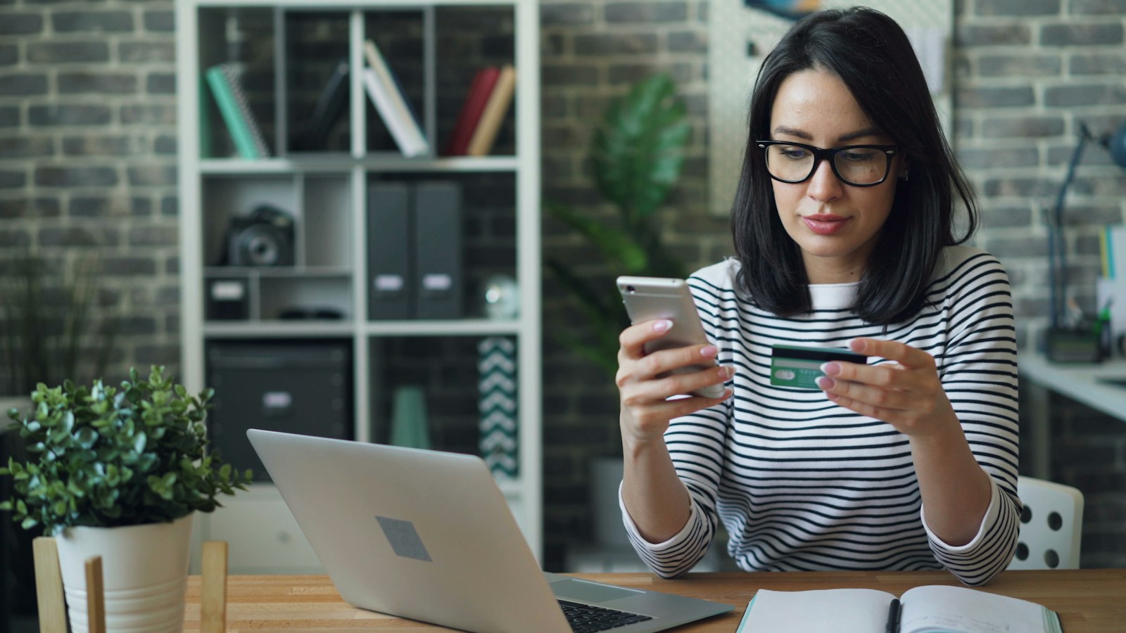 A woman in glasses and a striped top holds a phone and a payment card beside a laptop