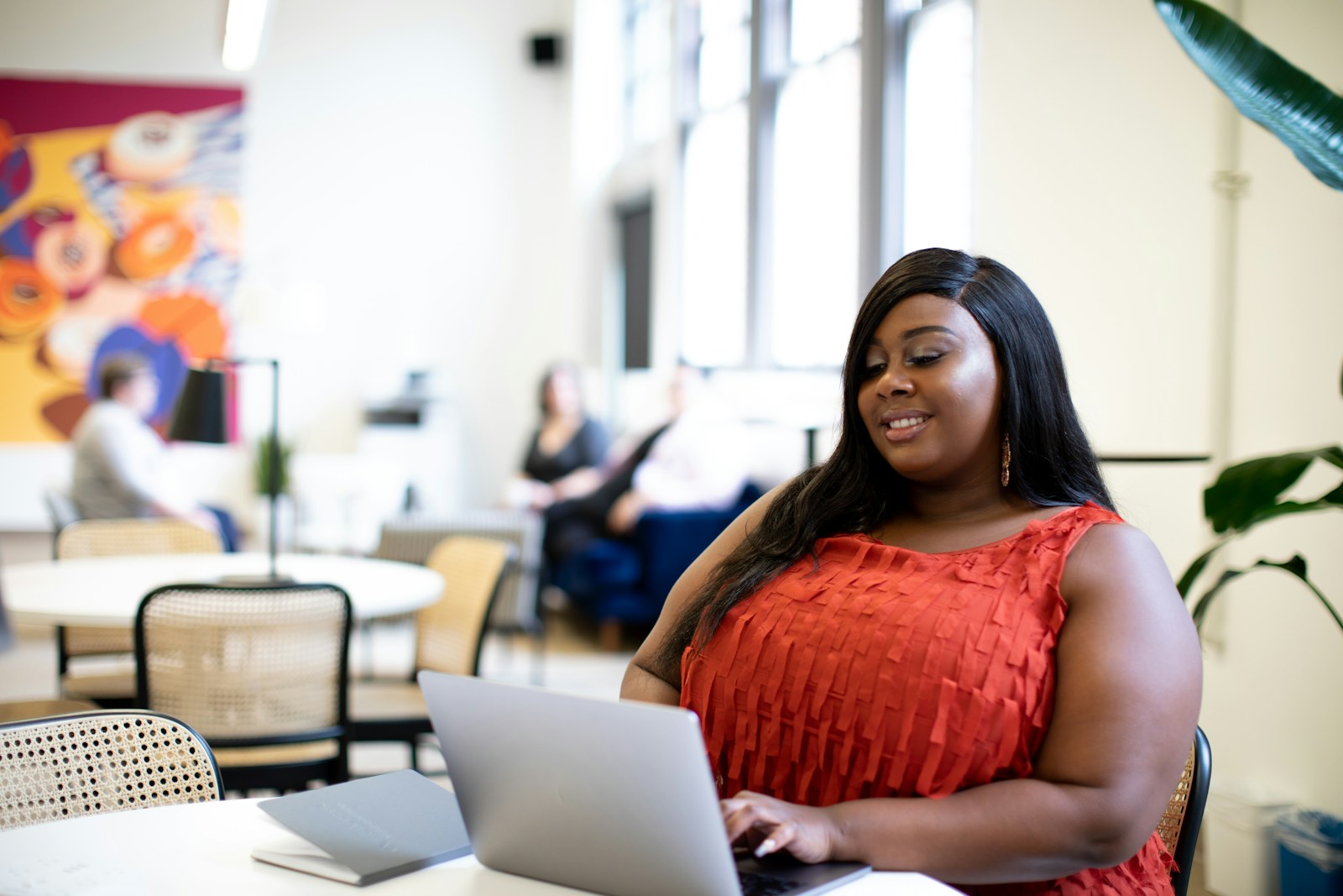 A woman in an orange top types on a silver laptop at a table in a bright shared workspace