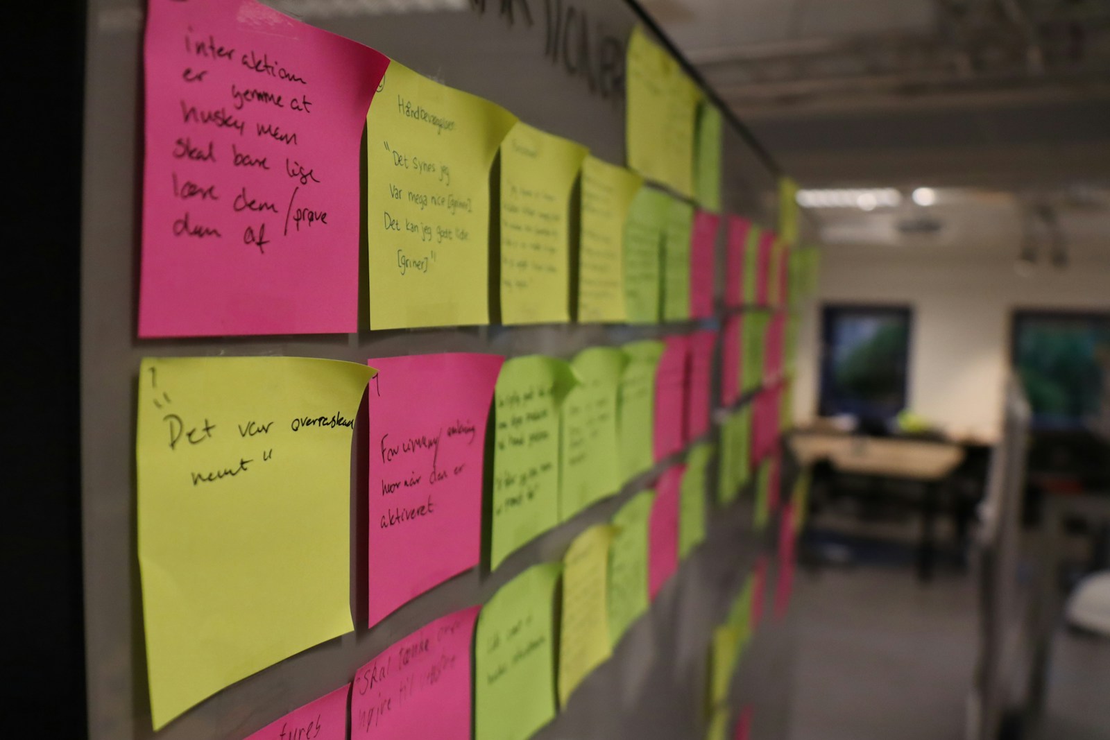 An office wall covered in rows of handwritten pink, yellow and green sticky notes, with a blurred meeting room behind