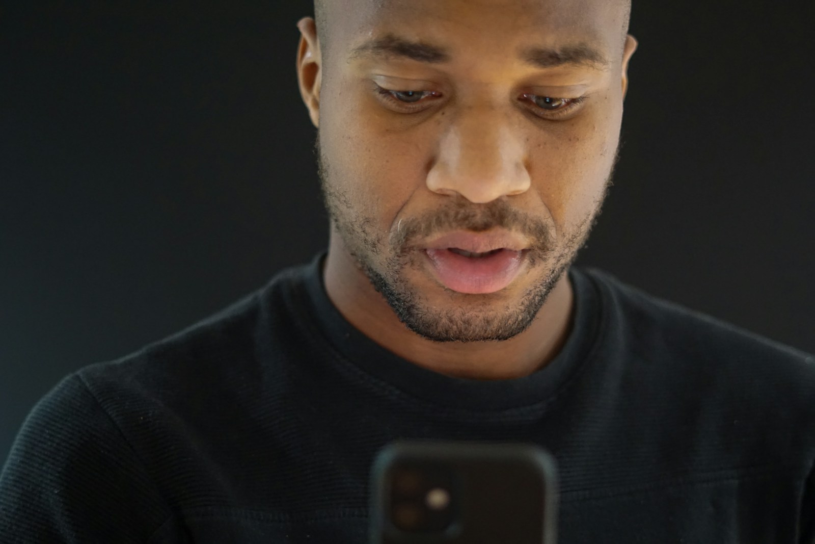 A man in a black sweater looks down at his smartphone against a dark background, mid-sentence