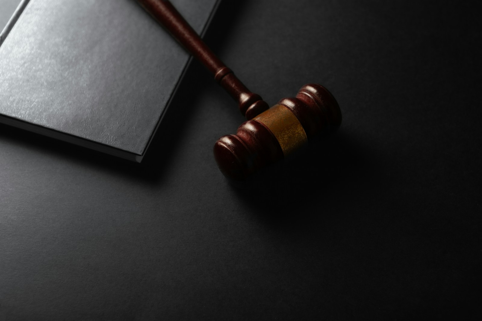 A wooden judge's gavel with a brass band resting beside a closed black book on a dark table
