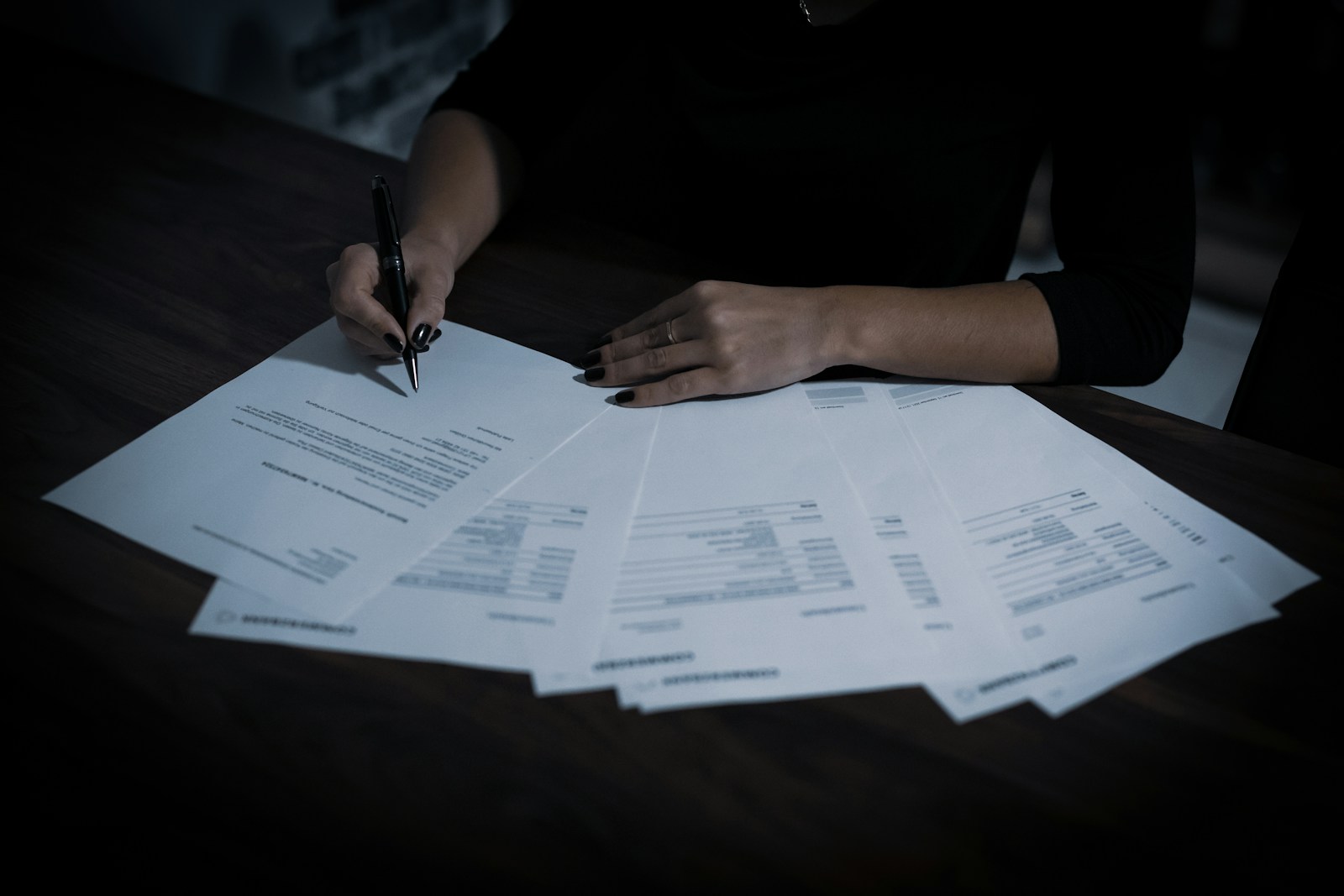 A woman marks one of several printed statements and forms fanned out across a dark wooden table, holding a black pen