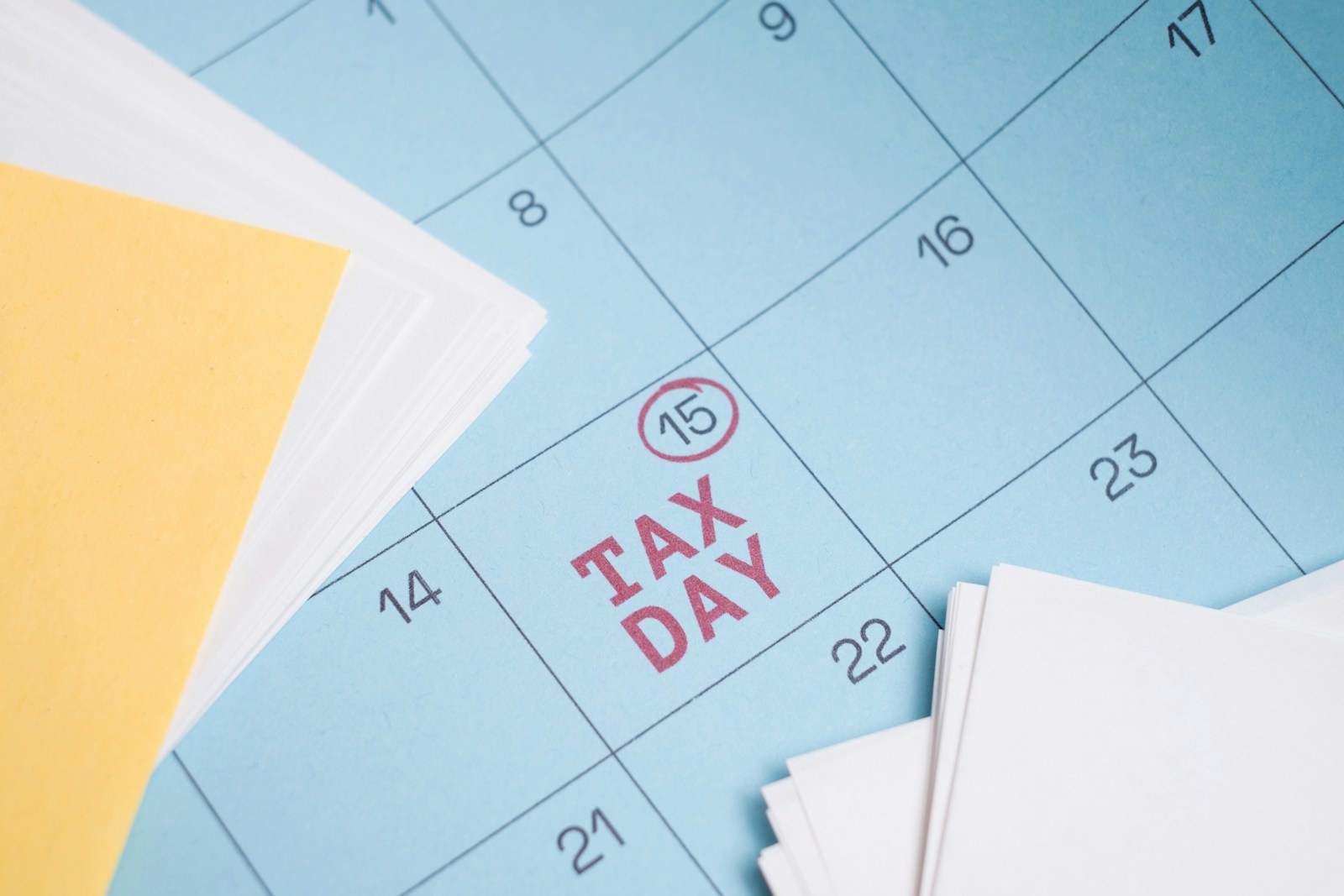 A blue calendar page with the 15th circled in red and Tax Day printed in its square, beside stacks of white and yellow paper