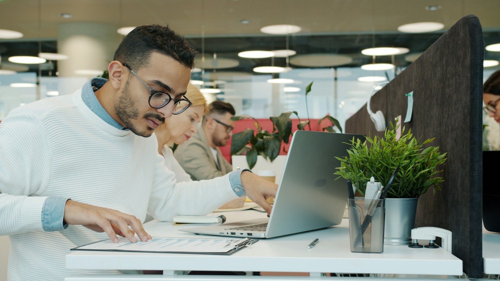 A man in glasses checks a document on a clipboard beside his laptop in a busy open-plan office