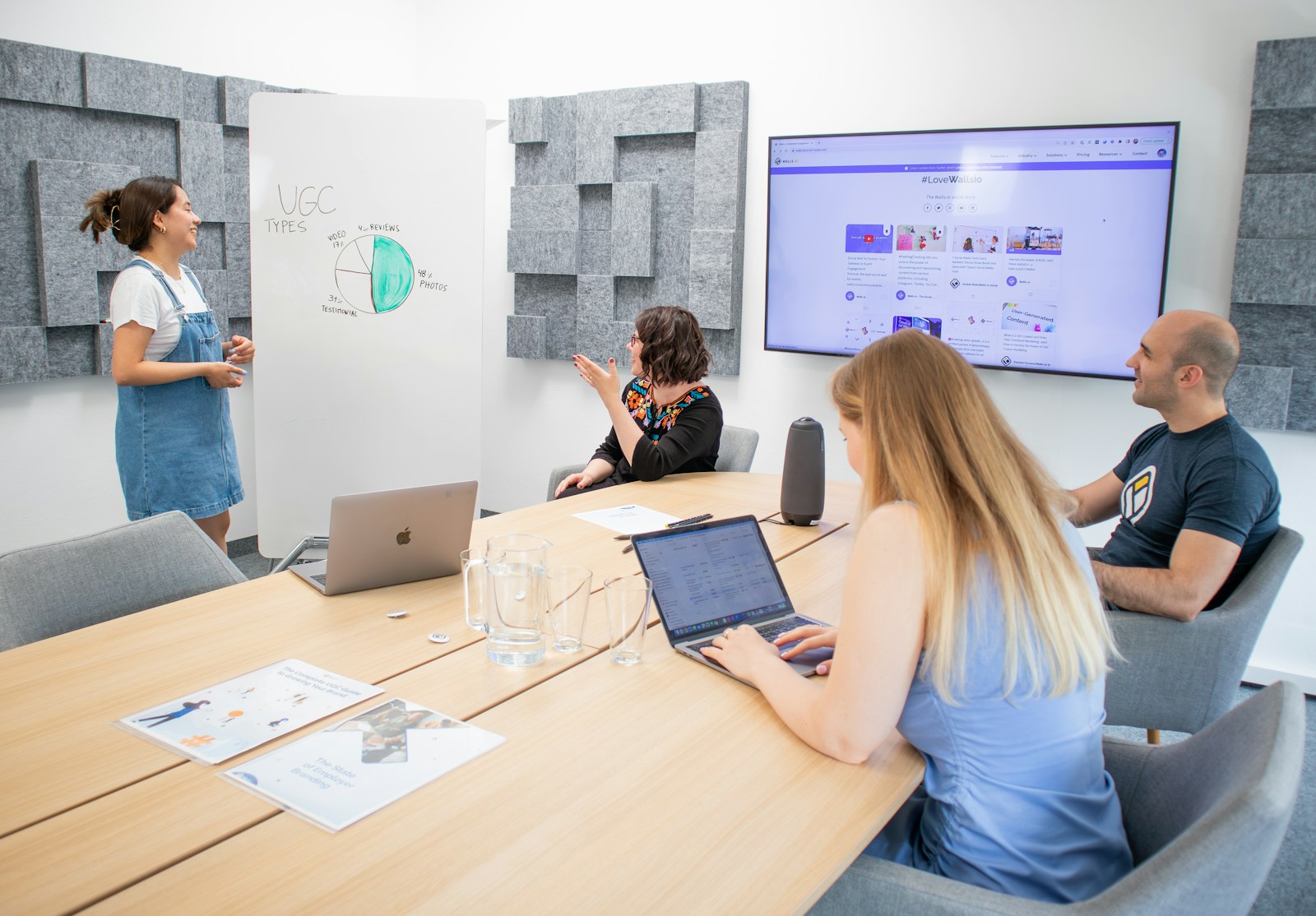 A woman in denim overalls presents a pie chart on a whiteboard to three colleagues at a meeting table with laptops