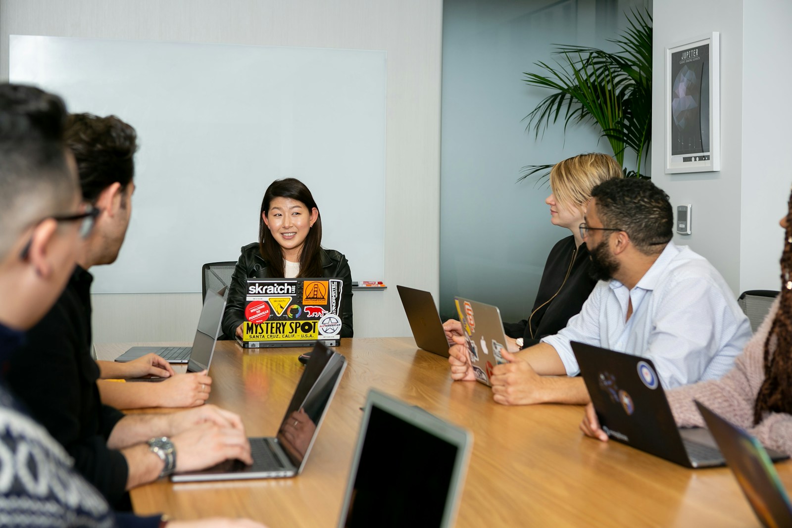 A woman at the head of a conference table talks to colleagues seated with open laptops along both sides