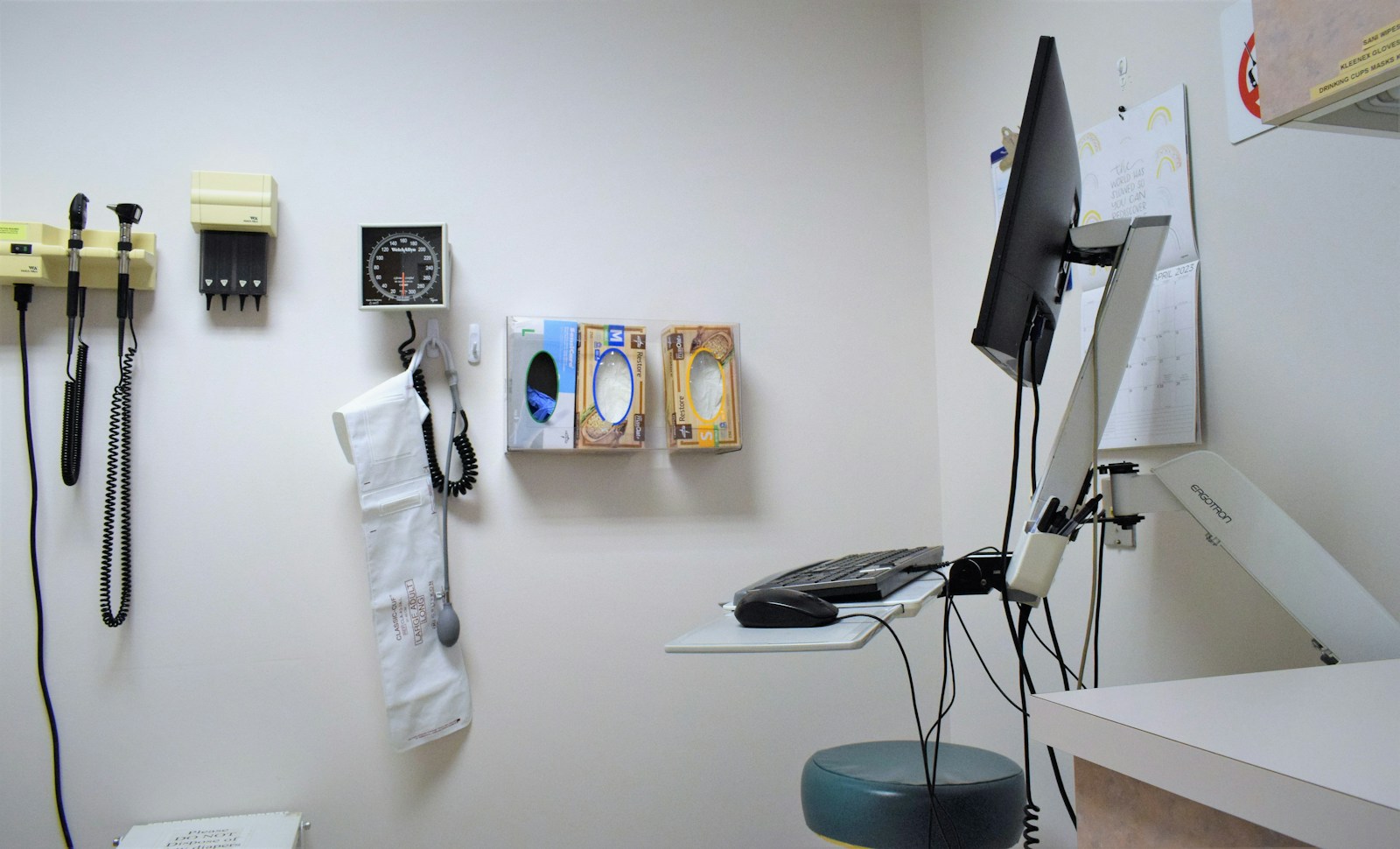 A medical exam room with wall-mounted diagnostic instruments, a blood pressure gauge, glove boxes and a computer workstation