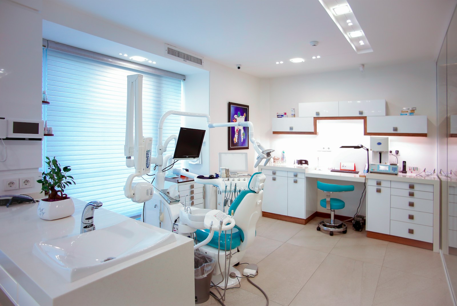 A bright dental treatment room with a teal patient chair, white cabinets, a monitor and window blinds