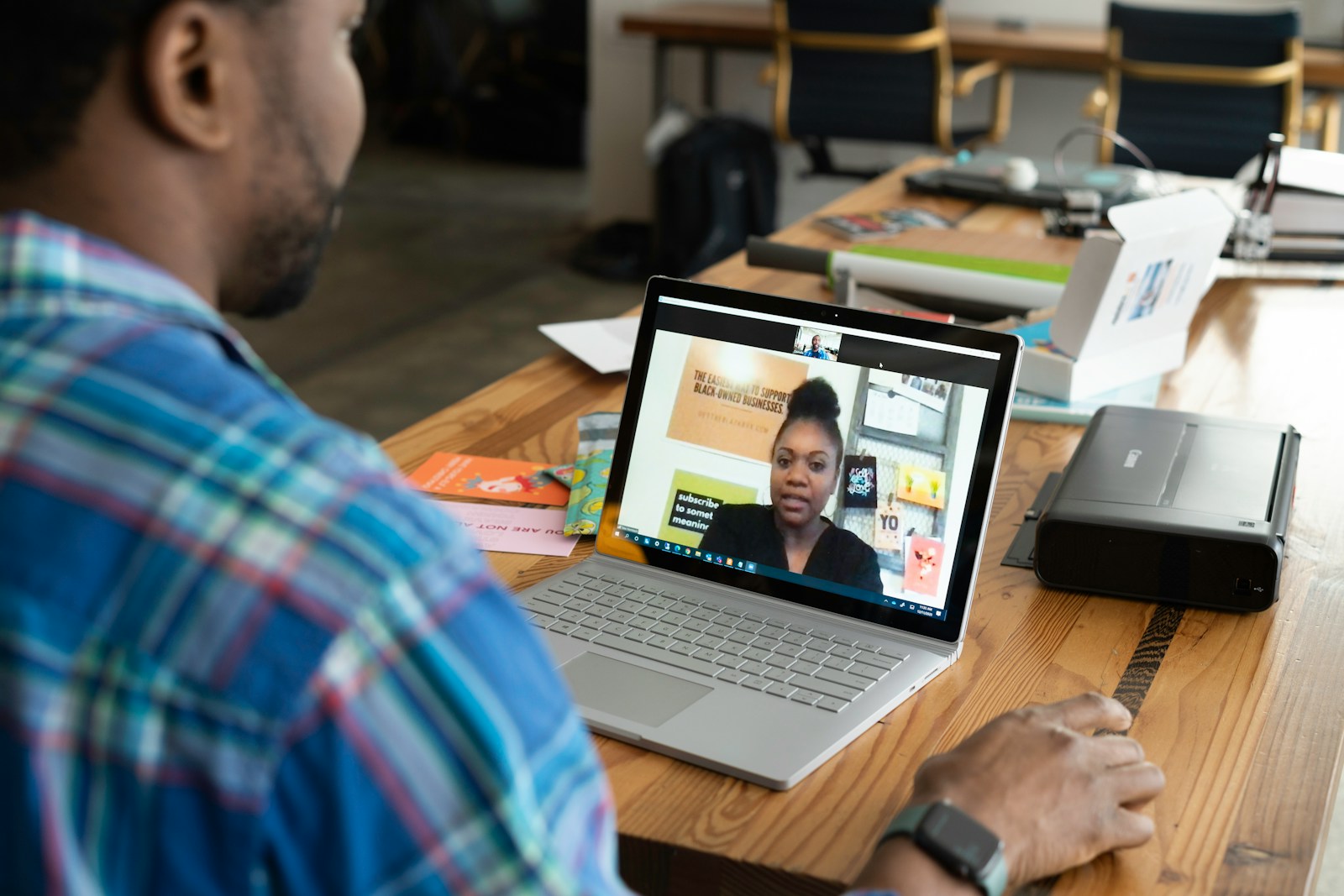 A man in a plaid shirt talks with a woman on a laptop video call at a wooden desk