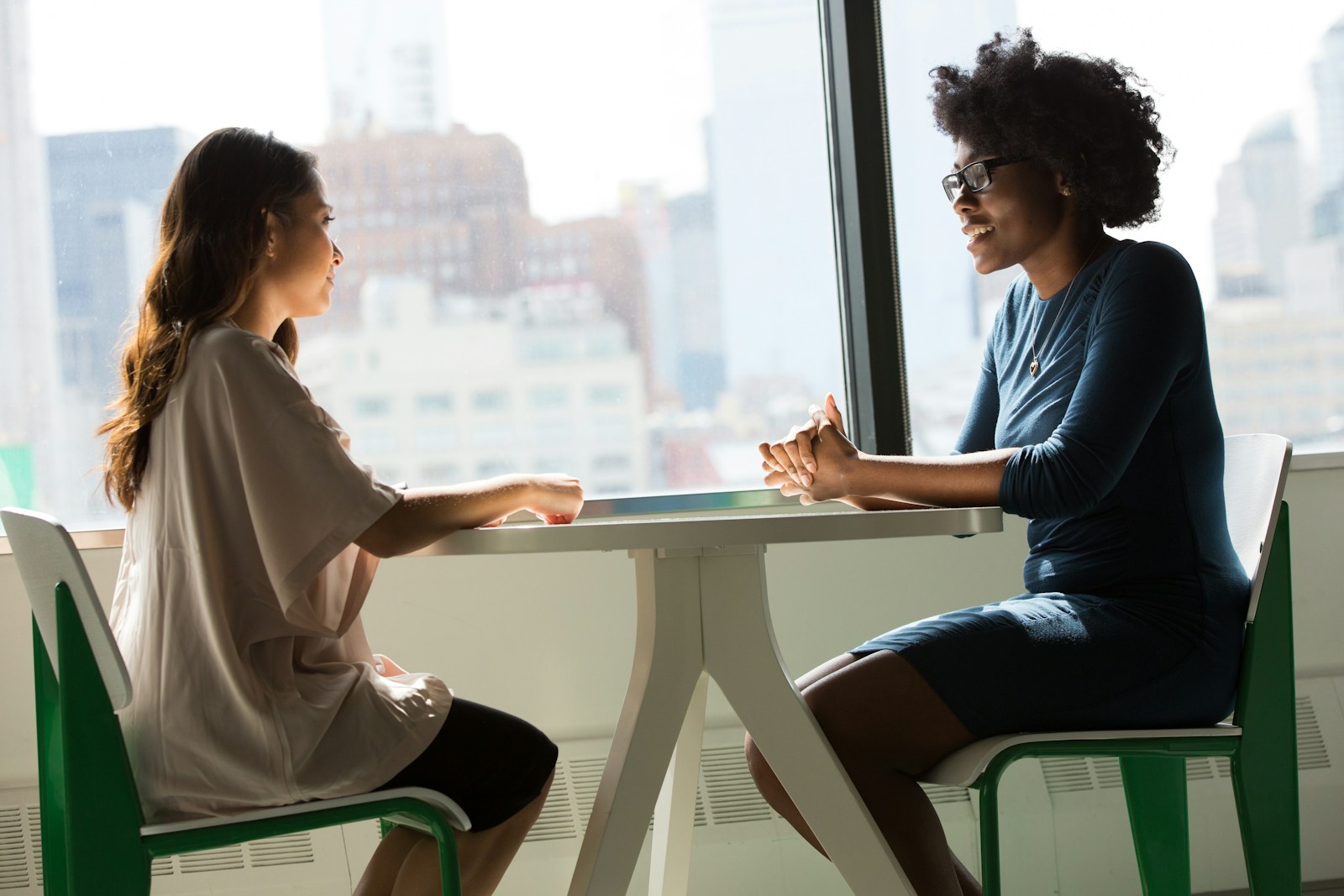 Two women sit facing each other at a small white table by a tall window, talking one to one