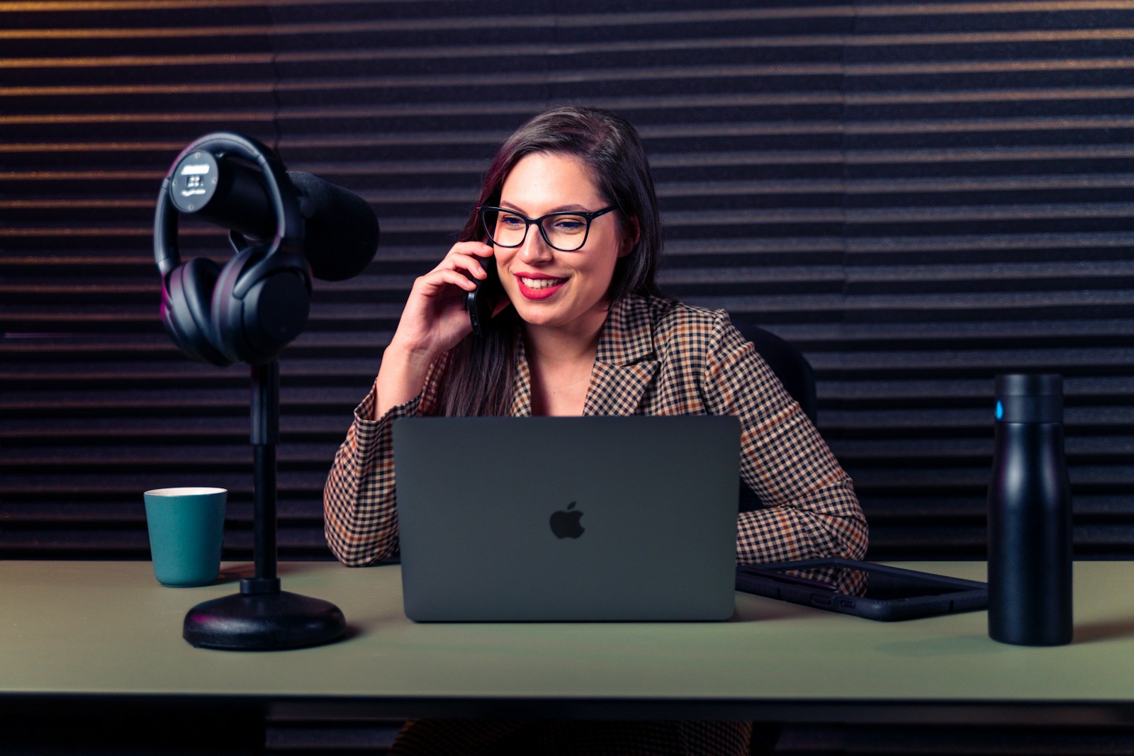 A smiling woman on a phone call works at a laptop beside headphones hanging on a desk microphone