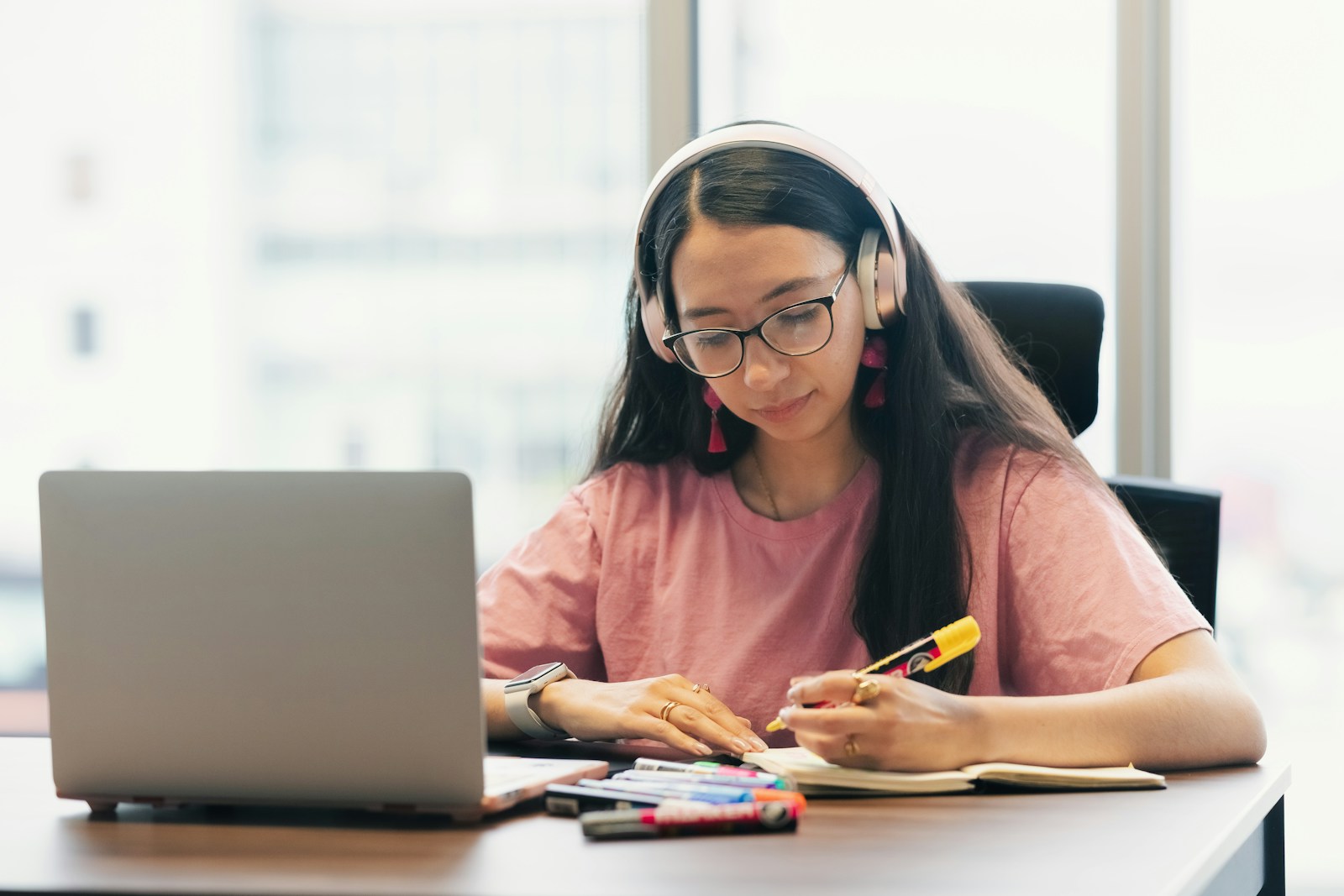 A young woman wearing headphones and glasses takes notes with a highlighter beside her open laptop