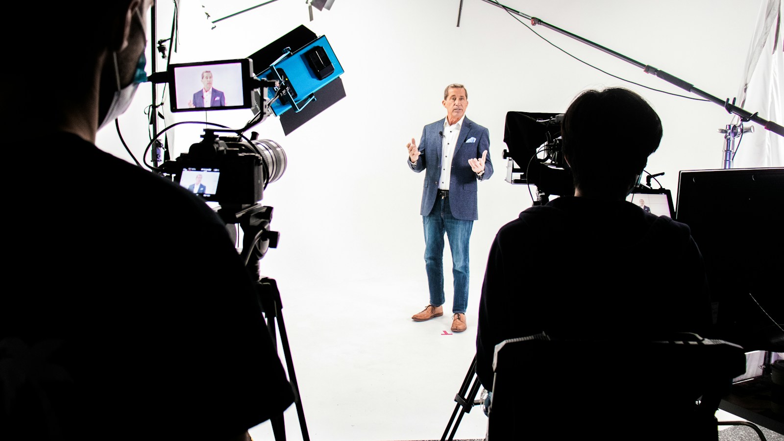 A presenter in a blue blazer talks to the camera in a white studio while two camera operators film him