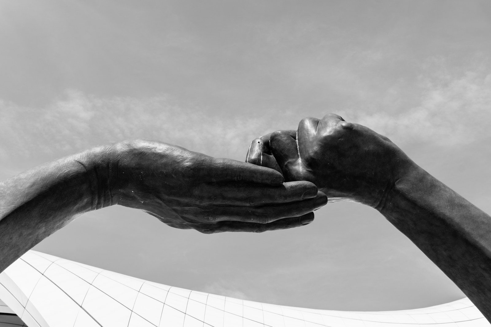 A black-and-white sculpture of two large hands meeting against a cloudy sky, one resting in the other