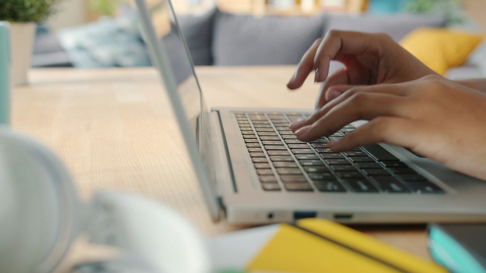 Hands typing on a silver laptop keyboard at a wooden table, with a yellow notebook blurred in the foreground