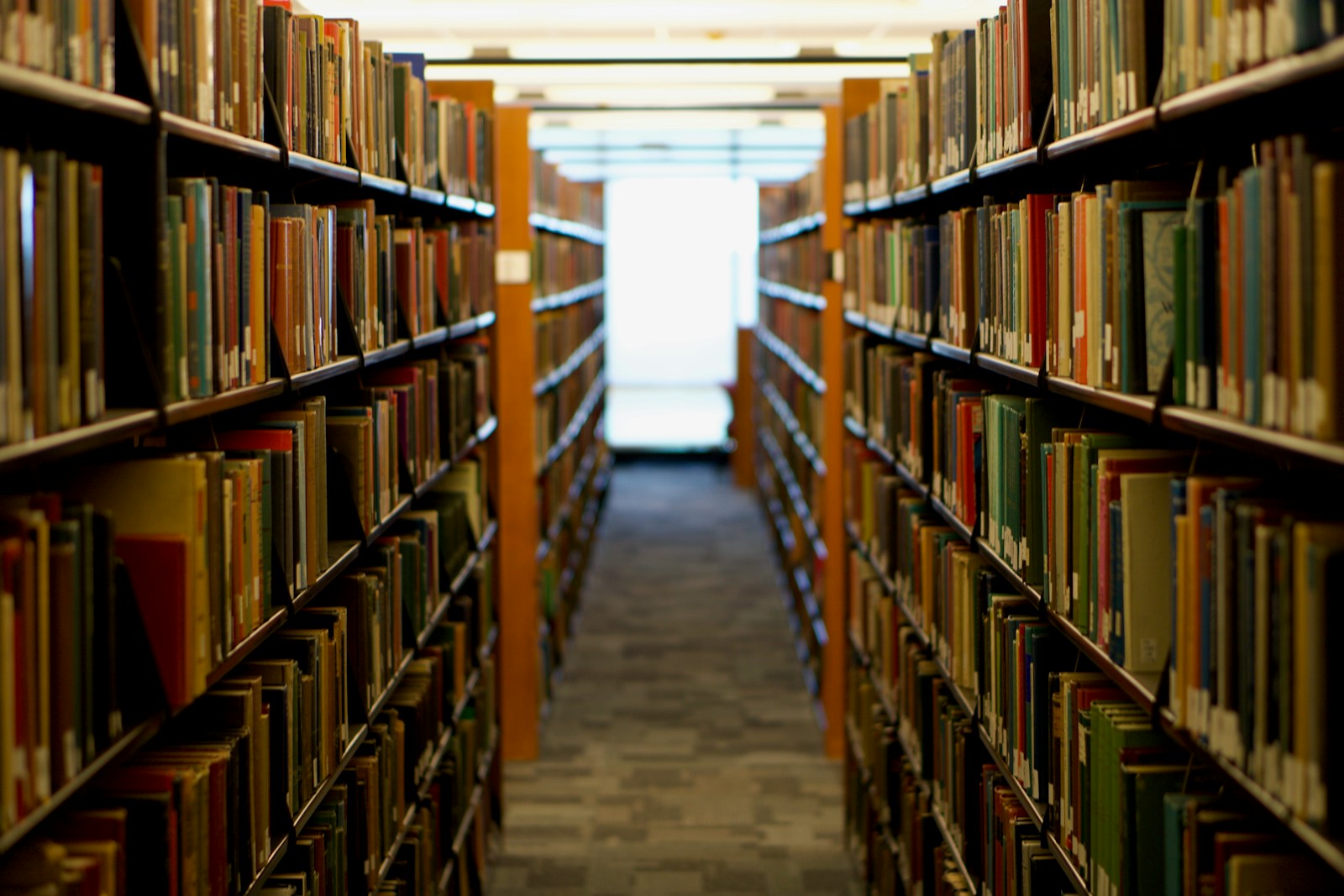 A long library aisle between tall shelves packed with books, lit by bright windows at the far end