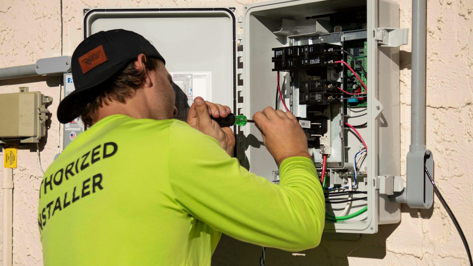 An electrician in a yellow installer shirt works with a screwdriver inside an open electrical panel fitted with circuit breakers
