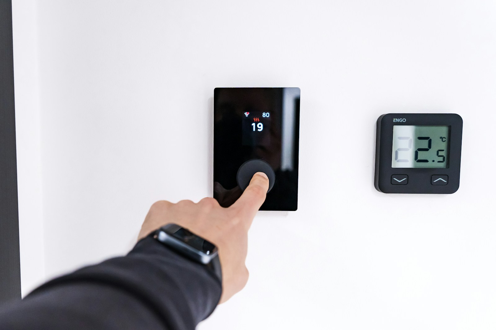 A hand presses the dial of a black wall thermostat showing 19 next to a digital thermometer reading 22.5 degrees