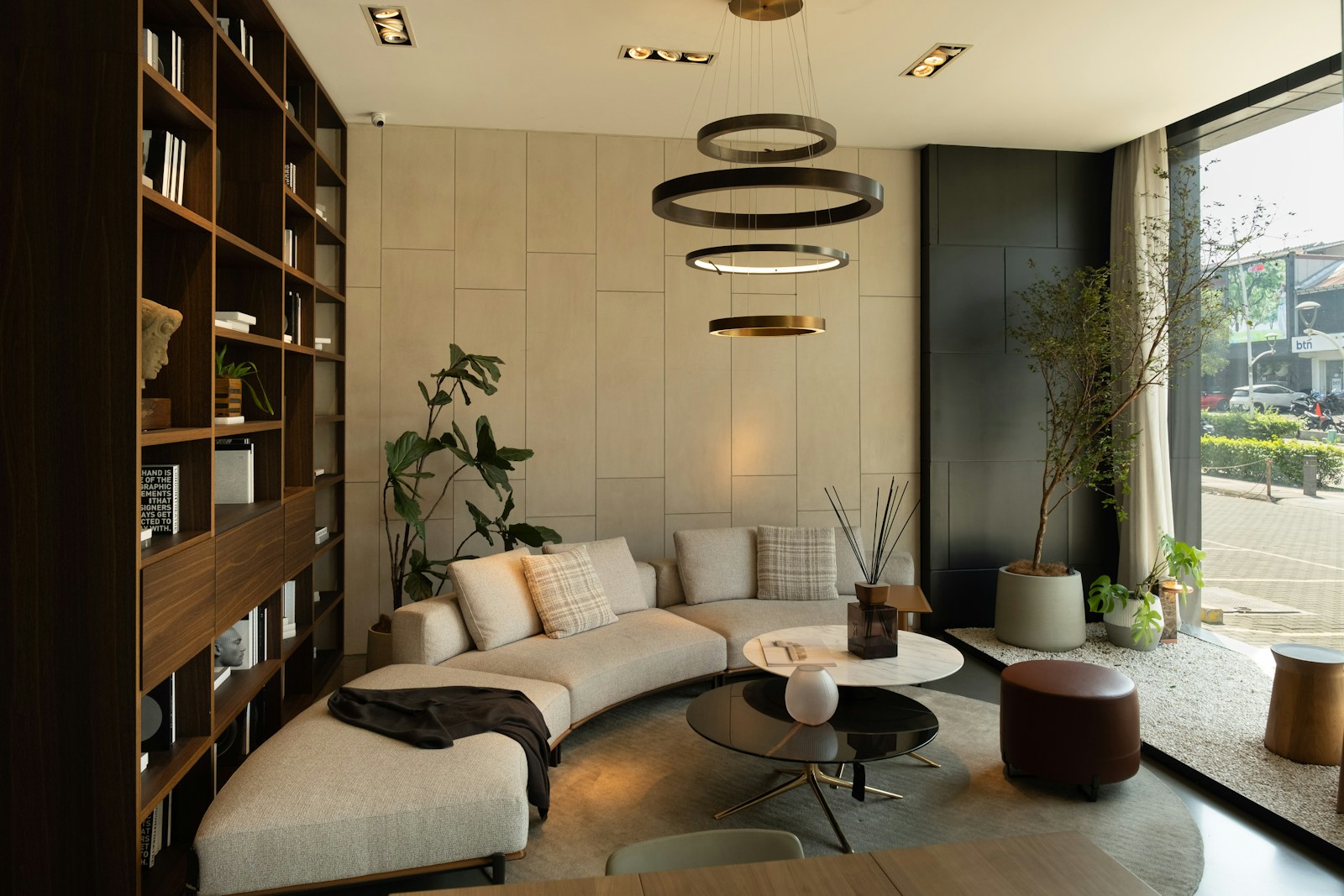 A modern living room with a curved beige sofa, round coffee tables, wooden bookshelves and a large street-facing window