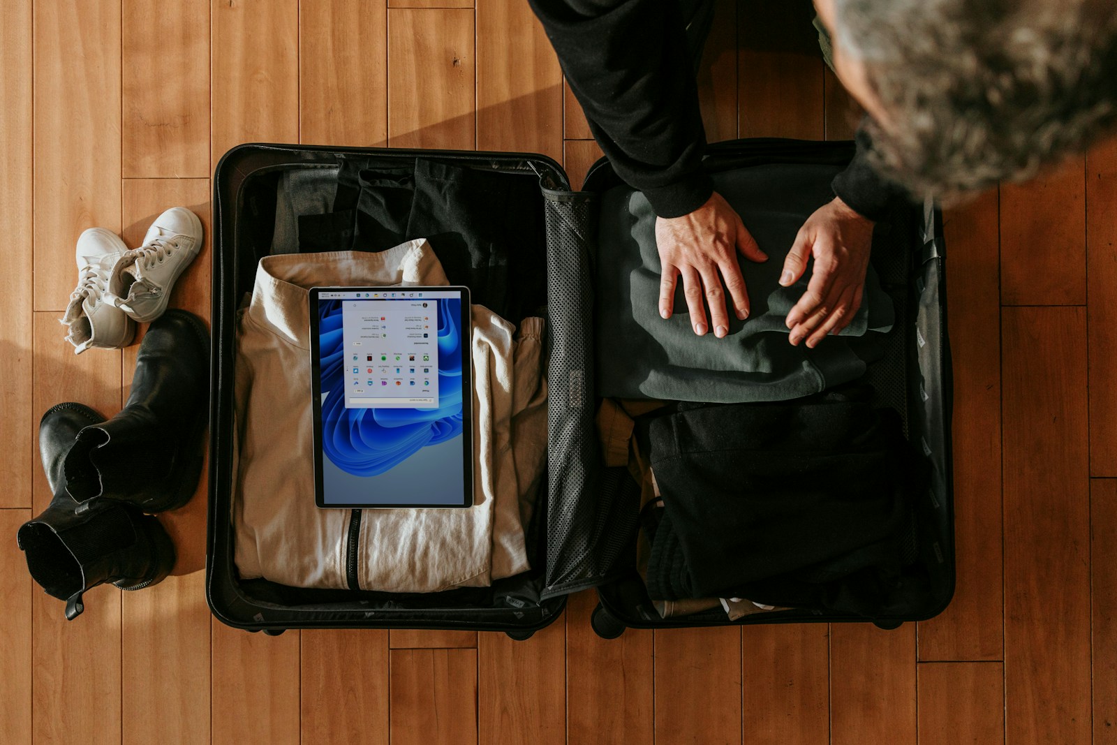 A person packs clothes into an open black suitcase on a wooden floor, a tablet resting on top