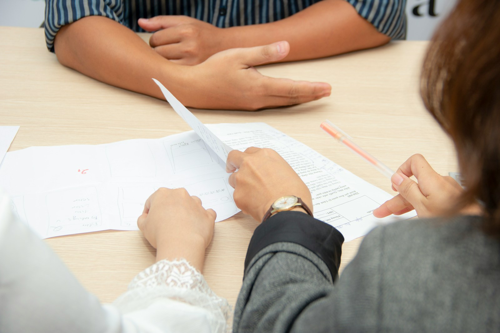 Two people review printed forms across a light wooden table, one holding a page and a pen during an intake interview