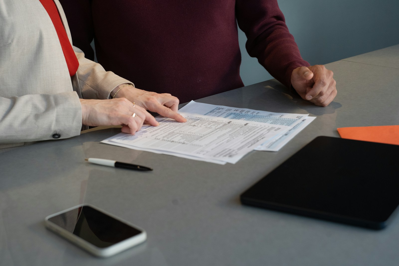 A woman in a cream blazer points to a line on a printed form while a man in a maroon sweater listens