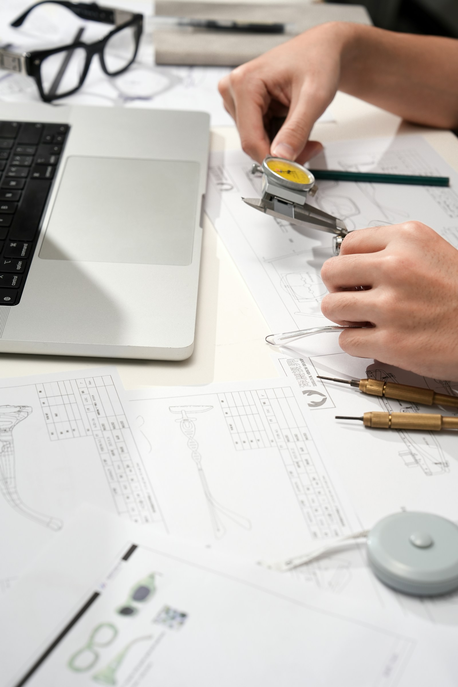 Hands measure a plastic eyeglass temple with a yellow dial caliper over technical drawings with dimension tables, beside a laptop