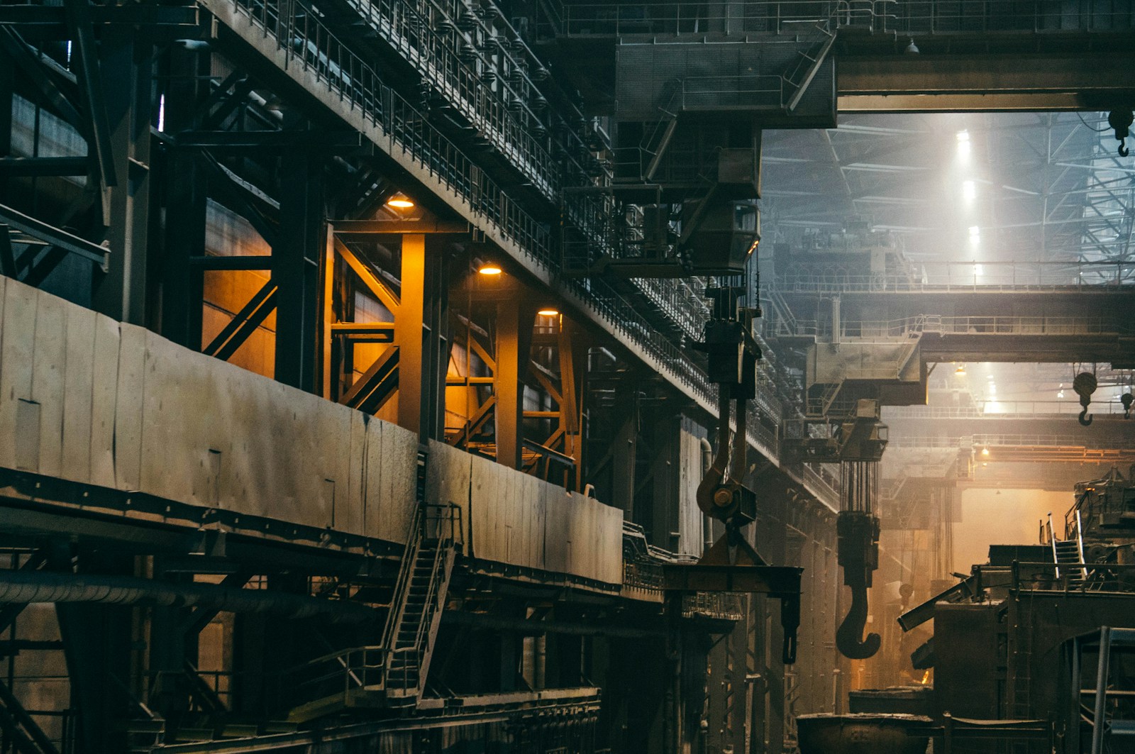 Overhead cranes with large hooks hang above a dim steel plant hall lit by orange lamps and skylights