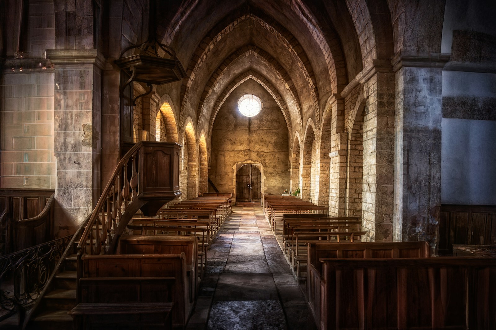 Empty wooden pews line the stone aisle of an old church with arched vaults, a wooden pulpit and a round window