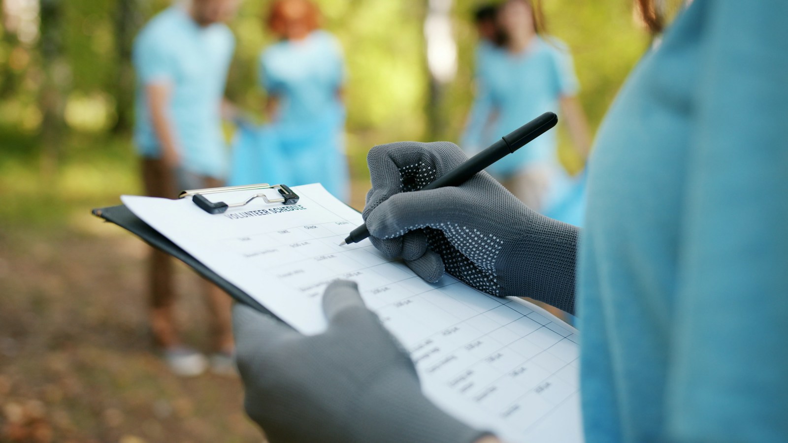A gloved hand writes on a clipboard headed Volunteer schedule while volunteers in light blue shirts stand blurred in the background