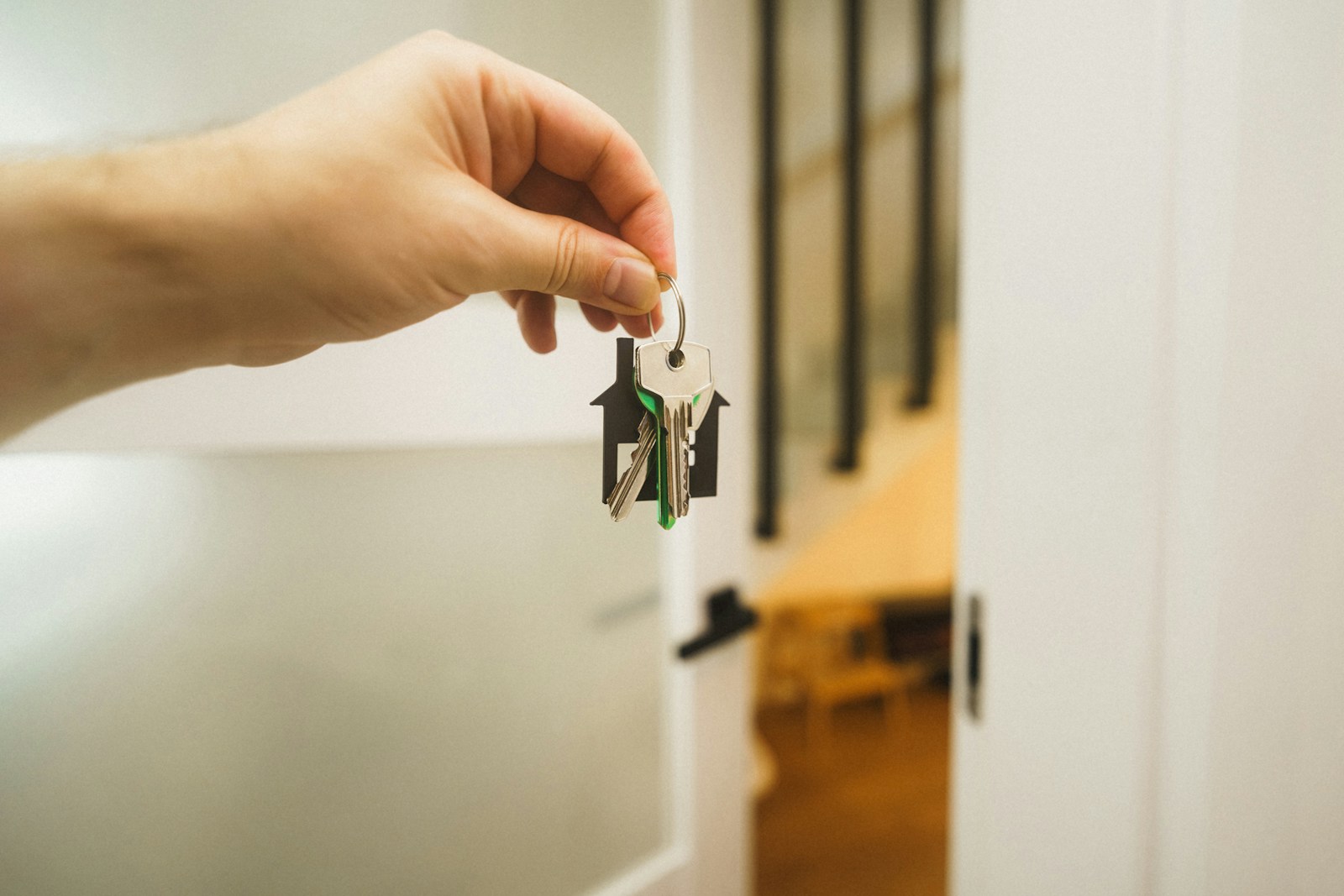 A hand holding a set of house keys on a house-shaped key ring in front of an open white door and a staircase