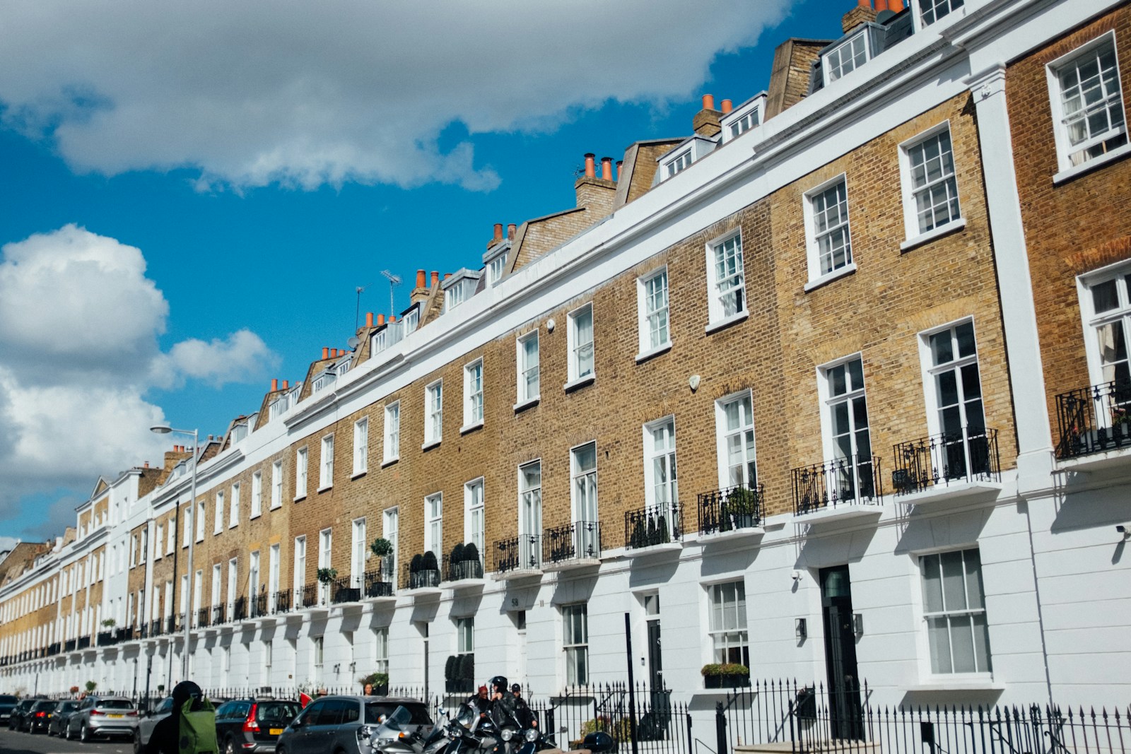 A row of brick and white terraced townhouses with black railings and parked cars on a sunny London street